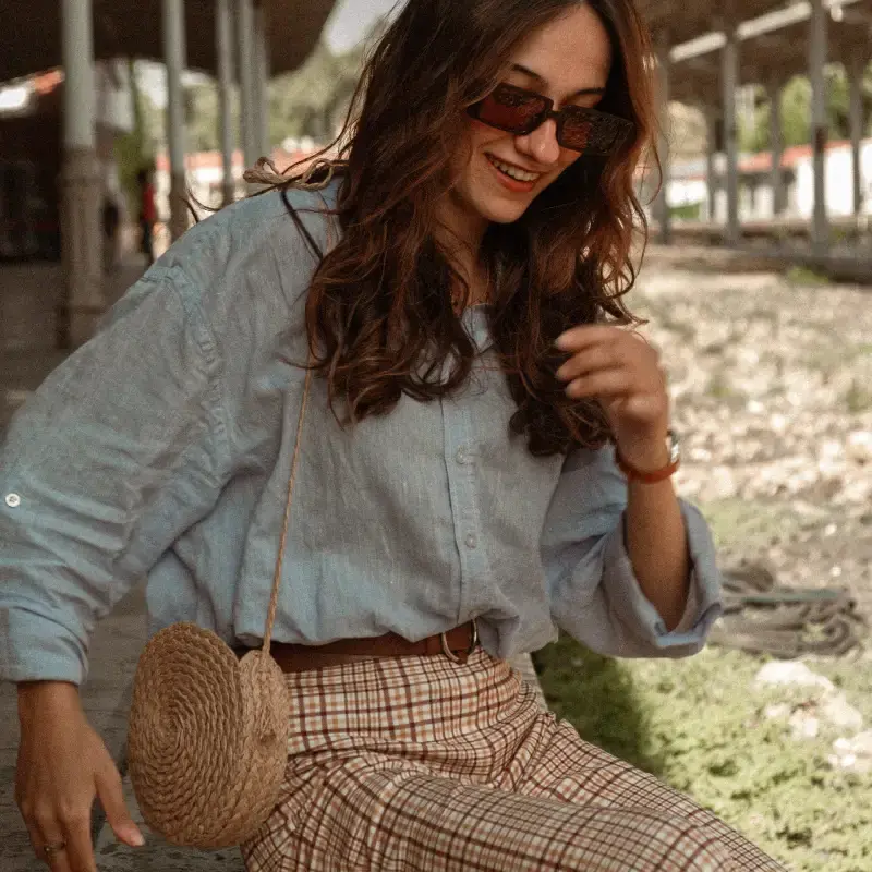 Mulher sentada em um banco na rua, vestindo calça xadrez e camisa e usando óculos escuros. Ela carrega uma bolsa de palha.